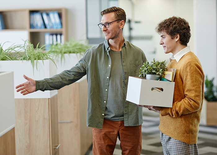 Two men in an office, one carrying a box, discussing work responsibilities and tasks. Two men in an office, one carrying a box, discussing work responsibilities and tasks.