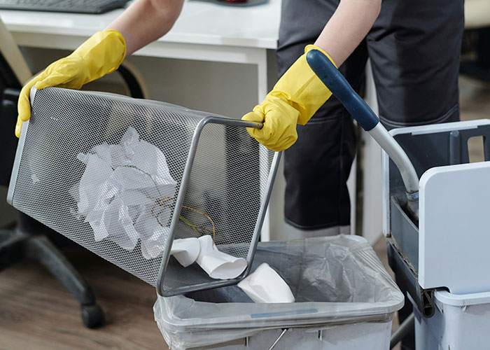 Worker cleaning office, emptying trash into bin with yellow gloves, emphasizing new guy's added workload. Worker cleaning office, emptying trash into bin with yellow gloves, emphasizing new guy's added workload.