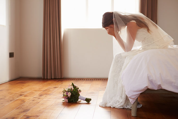 Bride sitting on a bed in distress, wedding bouquet on the floor; concept of wedding and family tension. Bride sitting on a bed in distress, wedding bouquet on the floor; concept of wedding and family tension.