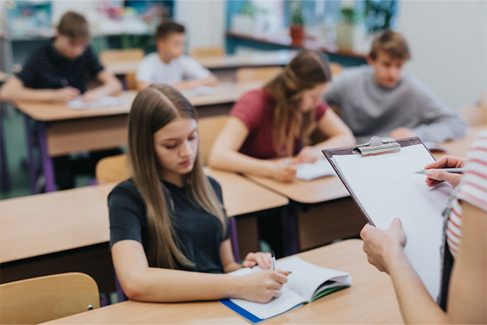 Student sits in class with a focused expression as teacher holds clipboard, reflecting on name pronunciation incident. Student sits in class with a focused expression as teacher holds clipboard, reflecting on name pronunciation incident.