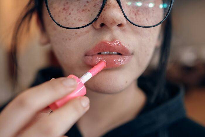 Person applying lip gloss, wearing oversized glasses, epitomizing recent fashion trends people hate.