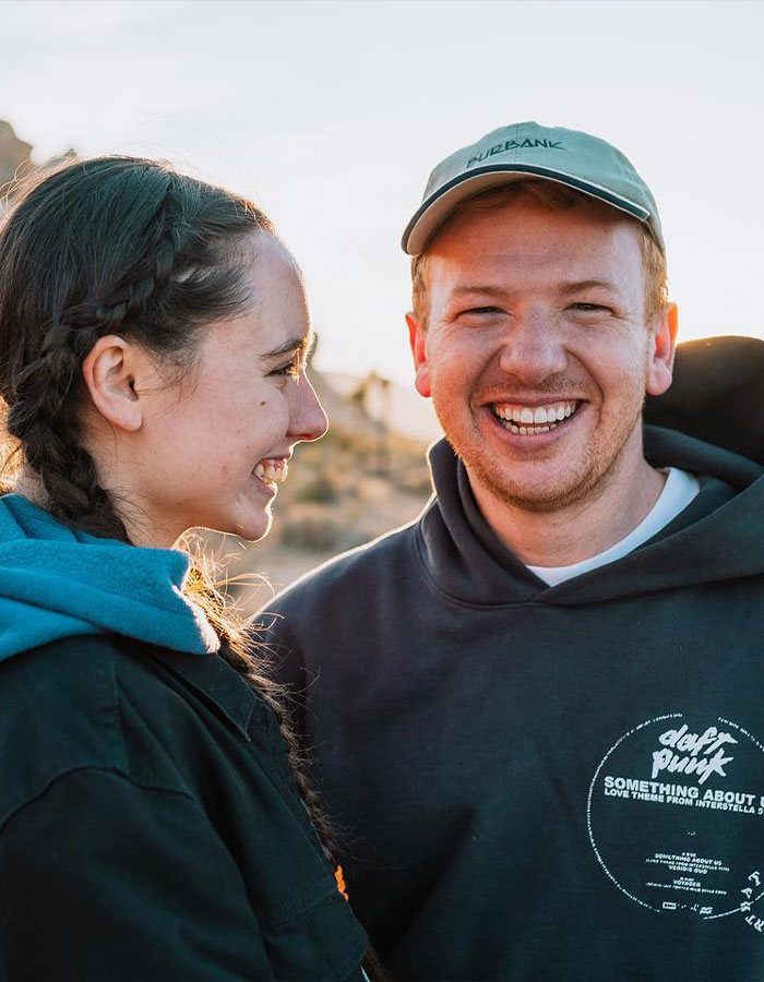 A couple smiling outdoors, woman in a braid and hoodie; man wearing cap and hoodie with Daft Punk logo. A couple smiling outdoors, woman in a braid and hoodie; man wearing cap and hoodie with Daft Punk logo.