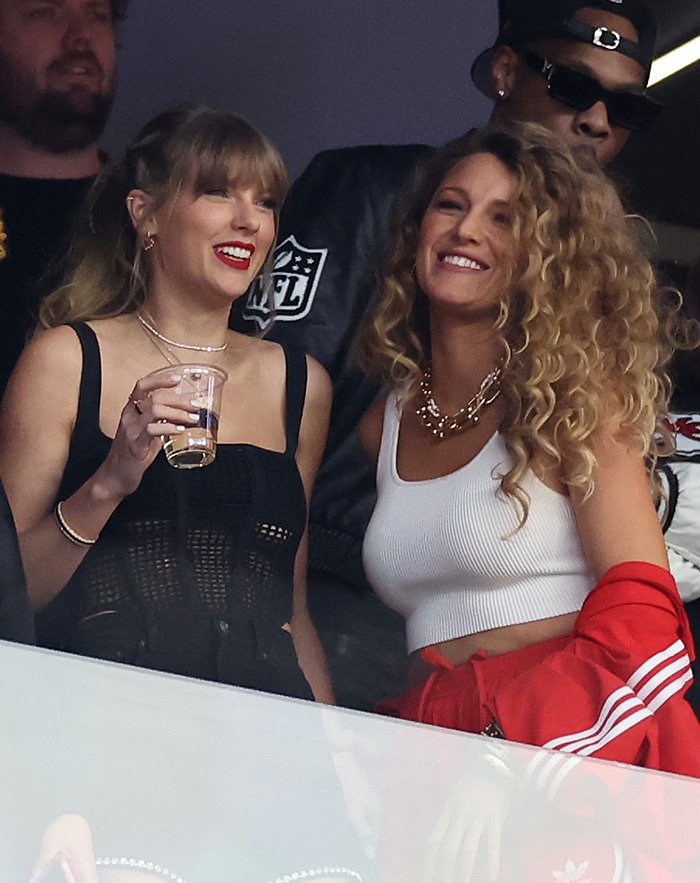 Taylor Swift and Blake Lively smiling at an event, one holding a drink, with NFL logo in the background. Taylor Swift and Blake Lively smiling at an event, one holding a drink, with NFL logo in the background.