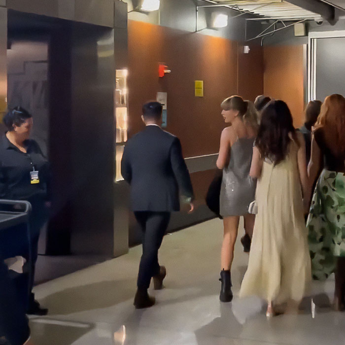 Taylor Swift leaving an event, wearing a silver dress, surrounded by people in an indoor hallway. Taylor Swift leaving an event, wearing a silver dress, surrounded by people in an indoor hallway.