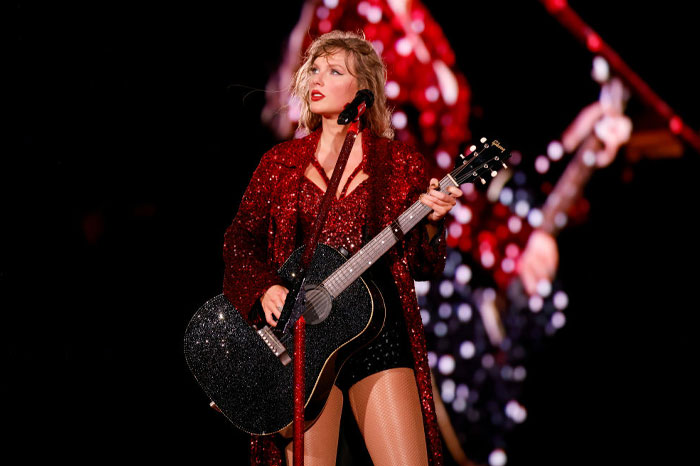 Taylor Swift performing on stage in a glittery red outfit, holding a black acoustic guitar. Taylor Swift performing on stage in a glittery red outfit, holding a black acoustic guitar.