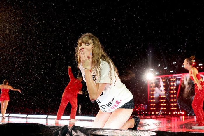 Taylor Swift performing on a wet stage, blowing a kiss, surrounded by dancers in red outfits. Taylor Swift performing on a wet stage, blowing a kiss, surrounded by dancers in red outfits.