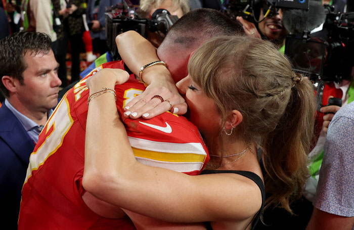 Taylor Swift hugging Travis Kelce in a stadium setting, surrounded by photographers.