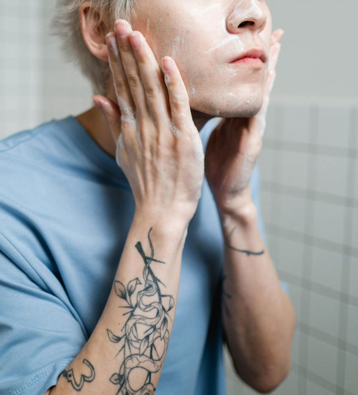 Teen applying skincare product, with tattoos visible on arms, in a tiled bathroom. Teen applying skincare product, with tattoos visible on arms, in a tiled bathroom.