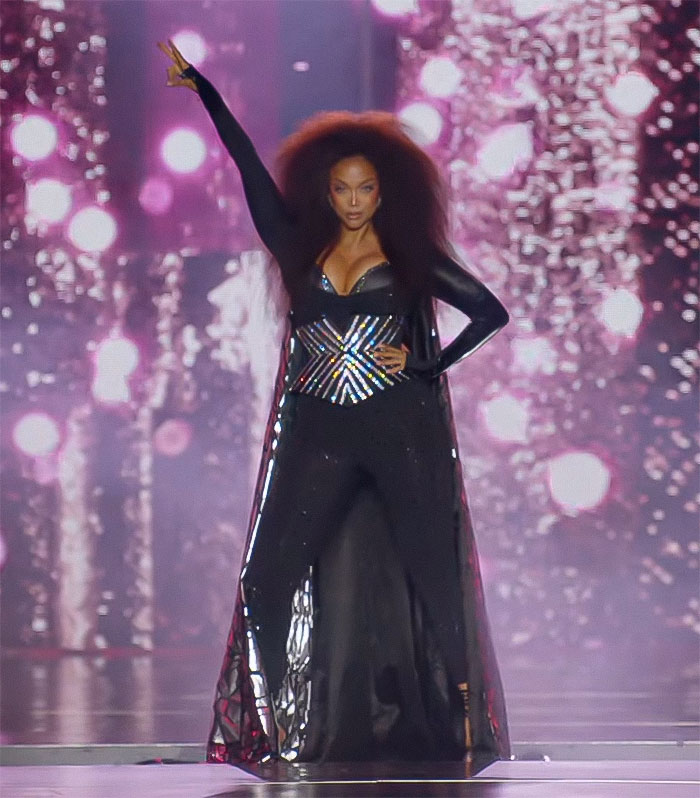 A person on stage in a black outfit, exuding confidence, against a backdrop of shimmering lights. A person on stage in a black outfit, exuding confidence, against a backdrop of shimmering lights.