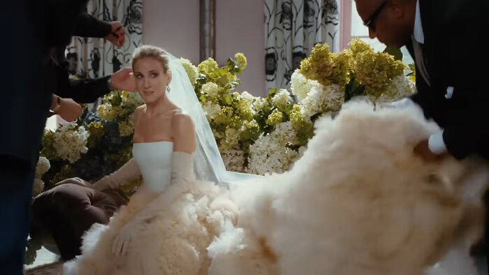 Woman in an extravagant wedding dress and veil, surrounded by people adjusting her gown, set against floral decor. Woman in an extravagant wedding dress and veil, surrounded by people adjusting her gown, set against floral decor.