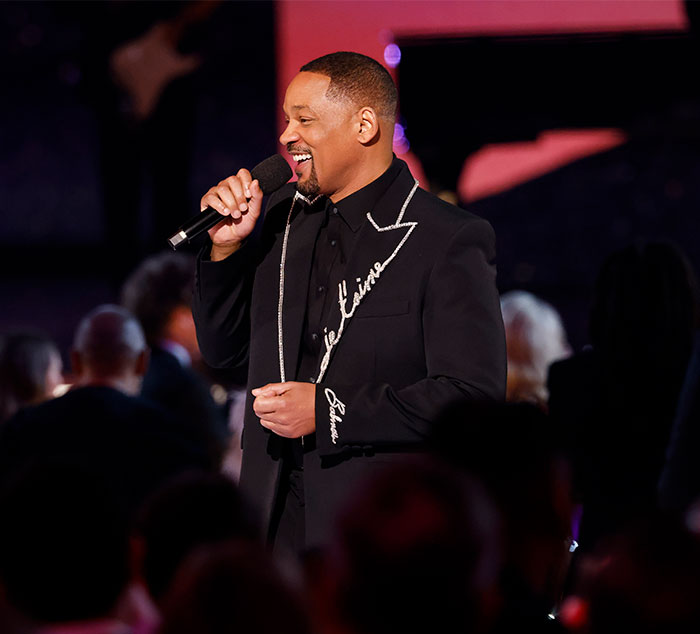 Man in formal attire holding a microphone on stage, associated with Grammy tribute and comeback event. Man in formal attire holding a microphone on stage, associated with Grammy tribute and comeback event.