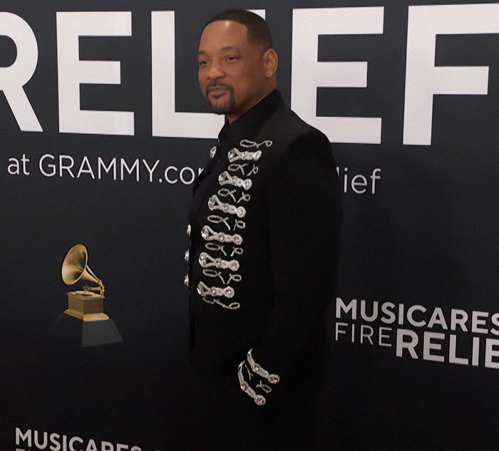 Man in a decorative black jacket stands beside a Grammy logo backdrop at an event. Man in a decorative black jacket stands beside a Grammy logo backdrop at an event.