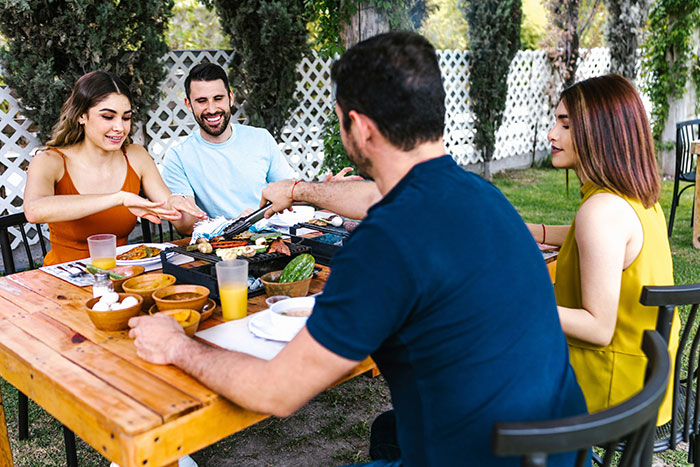 A group of friends enjoying a meal outdoors, laughing and sharing food at a table in a backyard setting. A group of friends enjoying a meal outdoors, laughing and sharing food at a table in a backyard setting.