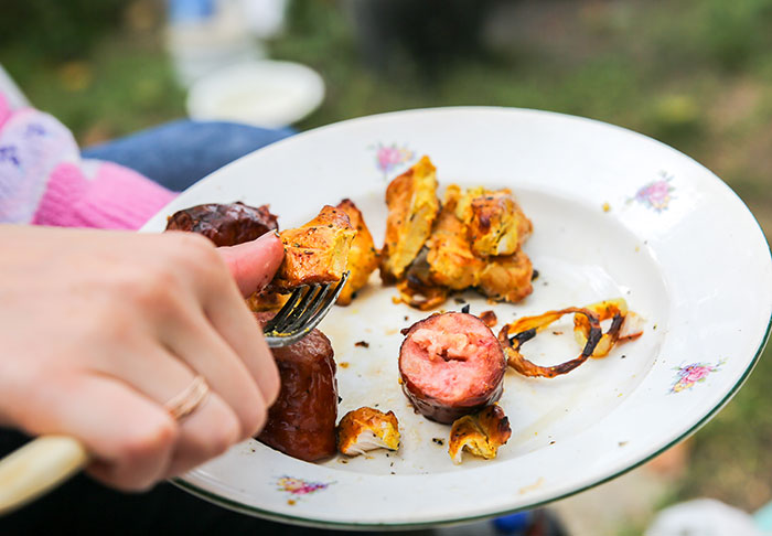 A person's hand holding a fork near a plate of leftovers, including grilled sausage and vegetables, at an outdoor gathering. A person's hand holding a fork near a plate of leftovers, including grilled sausage and vegetables, at an outdoor gathering.