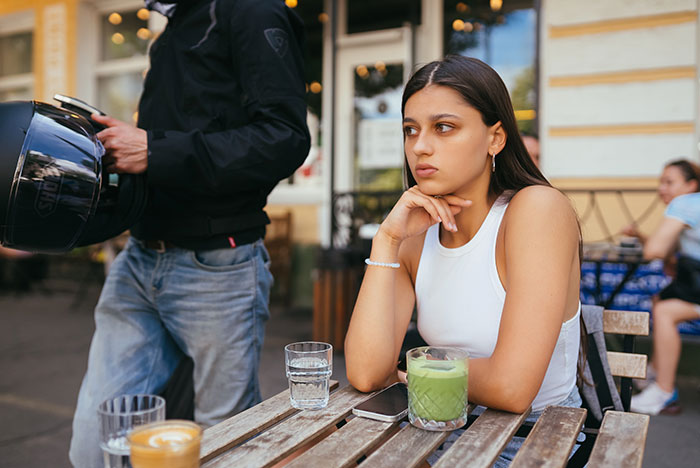 Woman at an outdoor cafe table with drinks, looking thoughtful, while a person stands nearby, not eating. Woman at an outdoor cafe table with drinks, looking thoughtful, while a person stands nearby, not eating.