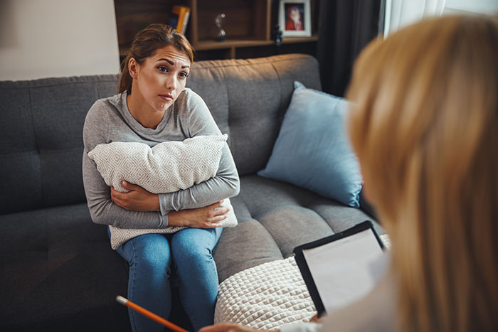 Woman on a couch, clutching a pillow, discussing adoption with another person in a home setting. Woman on a couch, clutching a pillow, discussing adoption with another person in a home setting.
