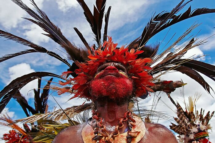 Person adorned in traditional attire with vibrant feathers and paint, representing global culture and tradition under a blue sky.