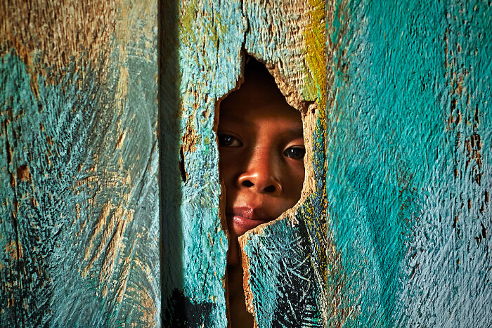 A child peeks through a hole in a textured wooden surface, capturing global culture and tradition.