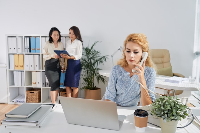 Office worker talking on phone, colleagues chatting in background; related to malicious compliance and egoistic boss. Office worker talking on phone, colleagues chatting in background; related to malicious compliance and egoistic boss.