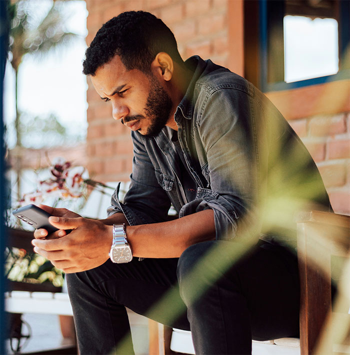 Man sitting on a porch, focused on his phone, surrounded by plants, wearing a denim shirt and silver watch. Man sitting on a porch, focused on his phone, surrounded by plants, wearing a denim shirt and silver watch.