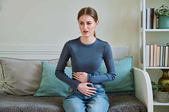 Person experiencing discomfort, holding stomach, possibly due to an allergy, sitting on a couch in a casual setting. Person experiencing discomfort, holding stomach, possibly due to an allergy, sitting on a couch in a casual setting.