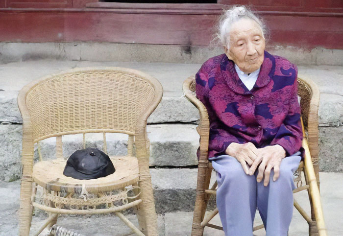 Elderly Chinese woman sitting outside on a wicker chair, wearing a purple jacket, with an empty chair beside her. Elderly Chinese woman sitting outside on a wicker chair, wearing a purple jacket, with an empty chair beside her.