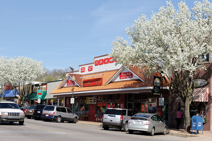 A street with parked cars and blooming trees near shops; a touristy place not worth the hype according to travelers.