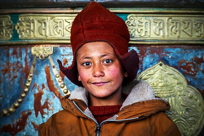 Child in traditional attire, wearing a red hat, in front of ornate cultural backdrop. Documenting culture and tradition.