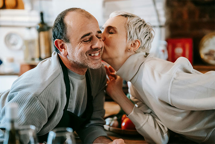 A woman in a cozy kitchen kisses a man wearing an apron, expressing familial affection. A woman in a cozy kitchen kisses a man wearing an apron, expressing familial affection.