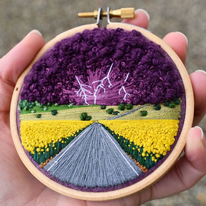 Hand holding a round embroidery featuring a vibrant landscape with a road and purple sky, crafted by Victoria Rose Richards.