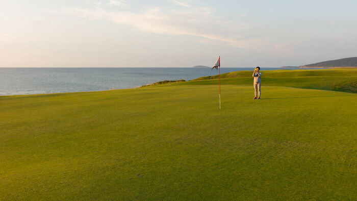 Golfer standing on a breathtaking course green, with picturesque ocean views and distant hills.
