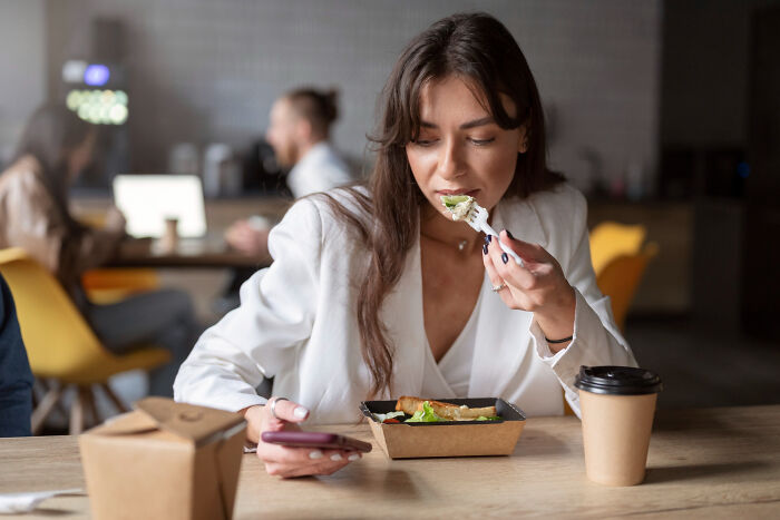 Woman eating salad at a cafe, smartphone in hand, surrounded by takeaway boxes—fun April Fools’ pranks theme.