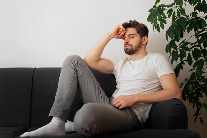 A pensive man in a white shirt and gray pants sits on a sofa, appearing thoughtful.