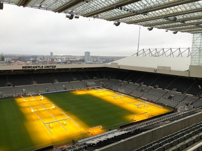 Soccer cathedral of St James' Park stadium with lighting system warming the green pitch under a cloudy sky.