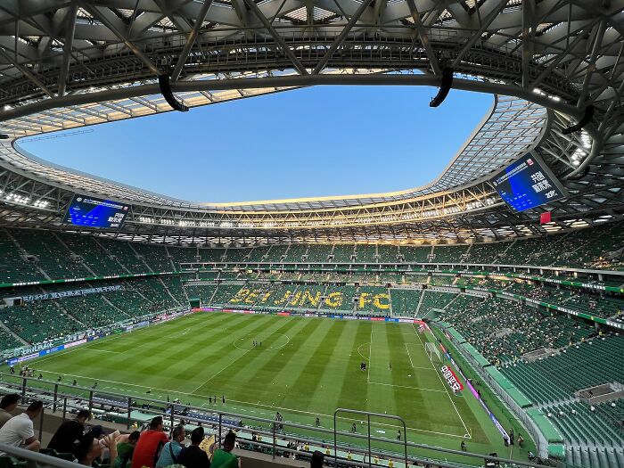 A modern soccer cathedral stadium with green seats and Beijing FC logo on the field during a sunny day.