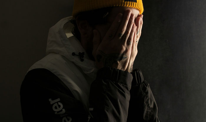 A person with a yellow beanie covers their face with hands, wearing a winter jacket, in a dark, moody setting. A person with a yellow beanie covers their face with hands, wearing a winter jacket, in a dark, moody setting.