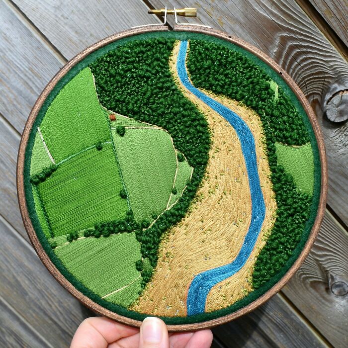 Landscape embroidery art with green fields and a blue river.