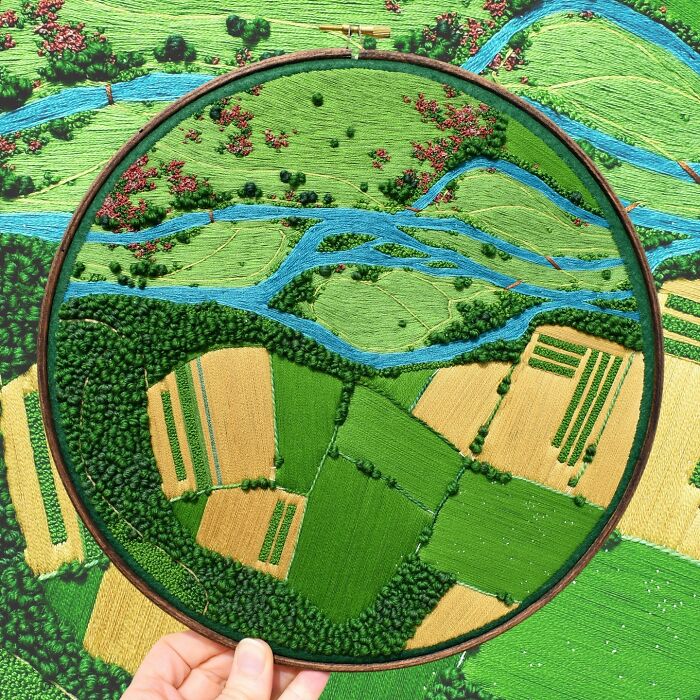 Hand holding a hoop displaying landscape embroidery with green fields and blue rivers by Victoria Rose Richards.