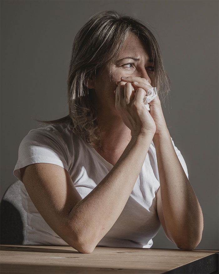A woman in a white shirt looking thoughtful, holding a tissue, with natural light highlighting her expression. A woman in a white shirt looking thoughtful, holding a tissue, with natural light highlighting her expression.