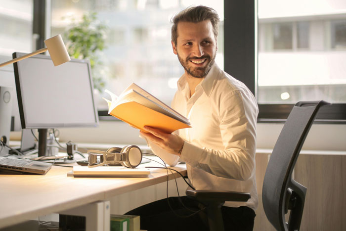 Man in a white shirt smiling at a desk, holding a yellow book, illustrating a calm reaction at work. Man in a white shirt smiling at a desk, holding a yellow book, illustrating a calm reaction at work.