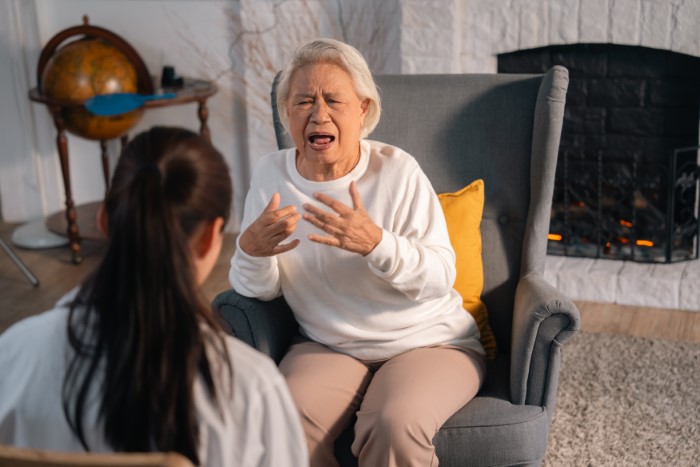 Grandmother in a white sweater gesturing emotionally while seated in a living room setting. Grandmother in a white sweater gesturing emotionally while seated in a living room setting.