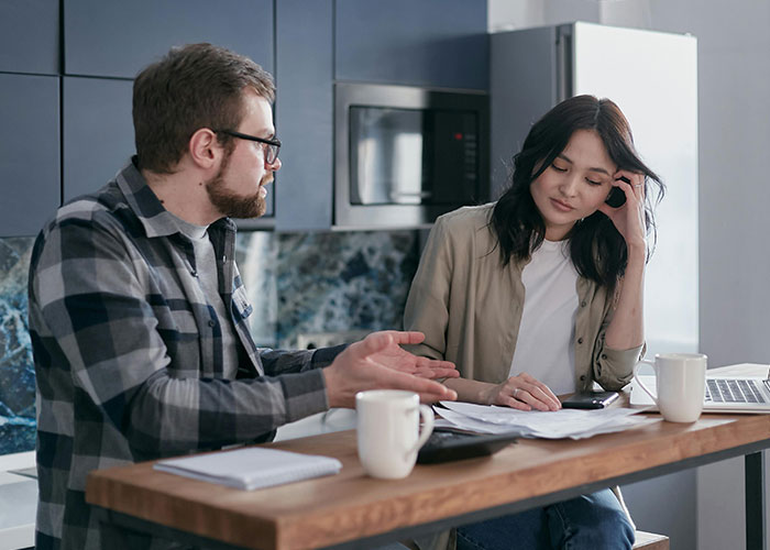 Man discussing property advice with woman at a kitchen table, surrounded by papers and coffee mugs. Man discussing property advice with woman at a kitchen table, surrounded by papers and coffee mugs.