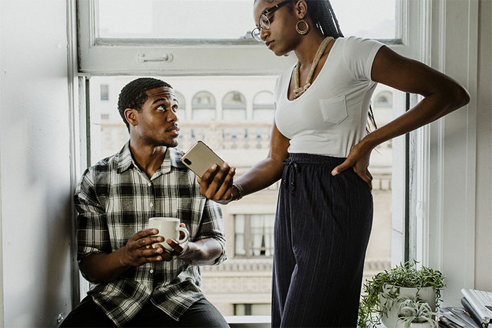 Girlfriend holding phone, tests boyfriend's loyalty; couple in a serious conversation by a window. Girlfriend holding phone, tests boyfriend's loyalty; couple in a serious conversation by a window.