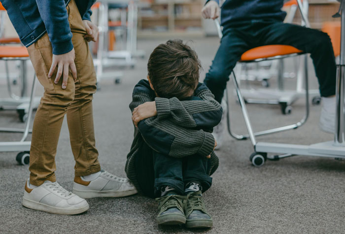 Young person sitting on the floor, hugging knees in distress, with two others nearby in a classroom. Young person sitting on the floor, hugging knees in distress, with two others nearby in a classroom.