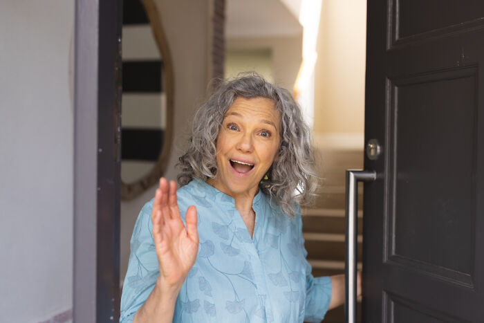 A woman with gray hair, wearing a blue shirt, joyfully waves from her open doorway; depicts insignificant thing moment.