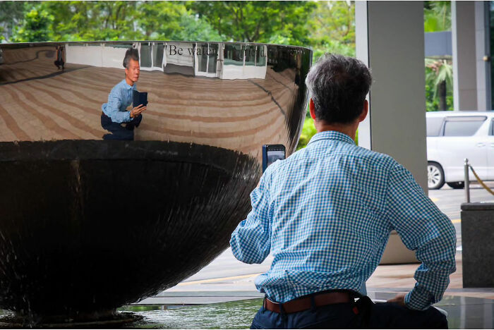 Photographer captures street life beauty with a reflection of a man in blue shirt holding a phone, mirrored in shiny surface.