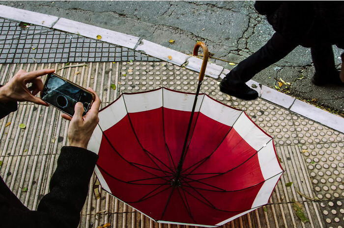 Street life photography: person capturing image of red umbrella on sidewalk with smartphone.