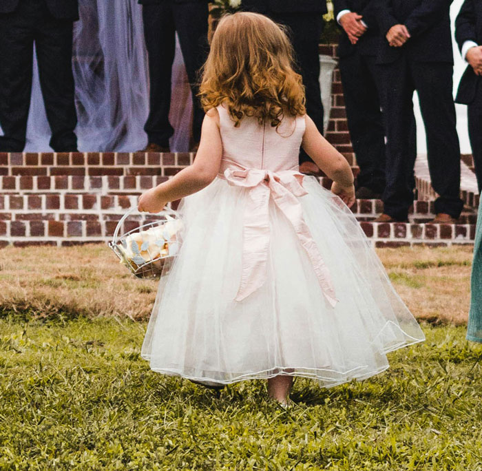 Flower girl in white dress holding basket, approaching wedding ceremony on grassy lawn. Flower girl in white dress holding basket, approaching wedding ceremony on grassy lawn.