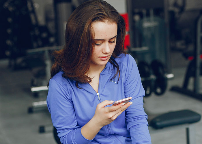 Woman in gym wearing blue jacket, focused on phone, surrounded by workout equipment. Woman in gym wearing blue jacket, focused on phone, surrounded by workout equipment.