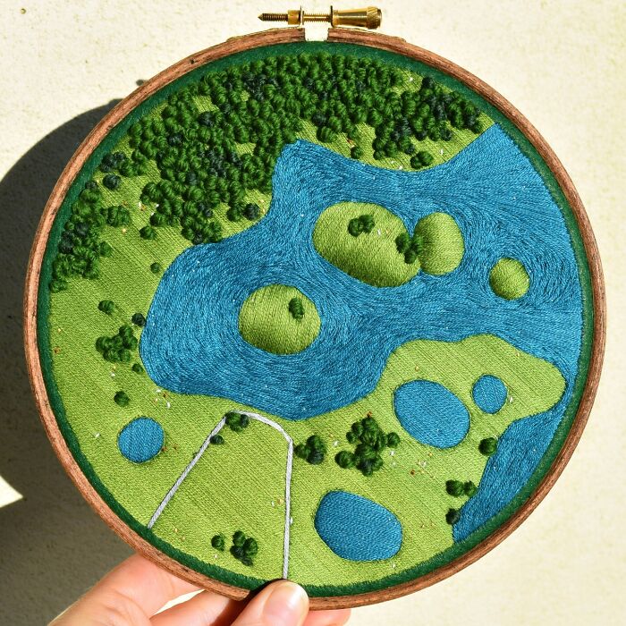 Landscape embroidery art with green fields, trees, and blue lakes by Victoria Rose Richards in a circular frame.